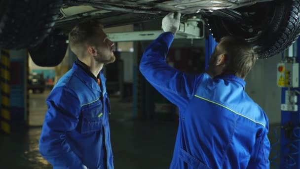 Jóvenes empleados del taller trabajando juntos debajo de un coche levantado — Vídeo de stock