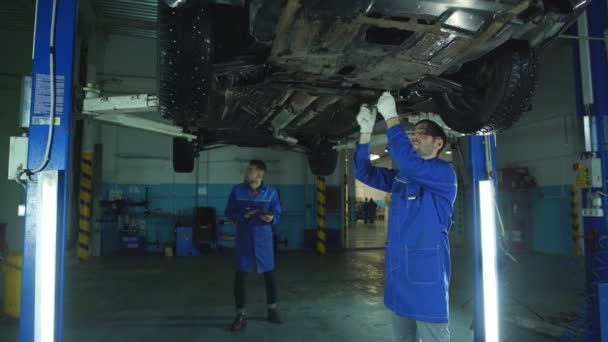 Mecánico y aprendiz trabajando debajo del coche — Vídeos de Stock