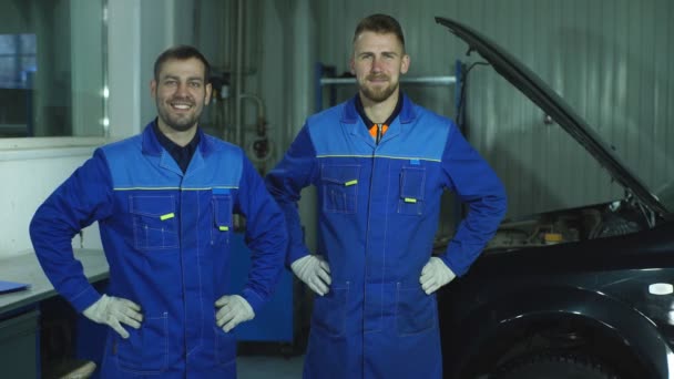 Vrolijke jonge mechanica gebaren terwijl voor de auto staan en lachend op camera — Stockvideo