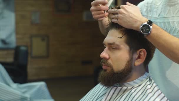 Joven barbudo hombre consiguiendo corte de pelo por peluquería mientras sentado en silla en barbería — Vídeo de stock