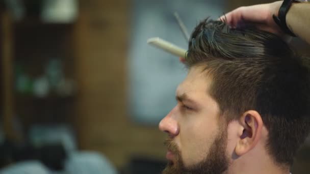 Joven barbudo hombre consiguiendo corte de pelo por peluquería mientras sentado en silla en barbería — Vídeos de Stock