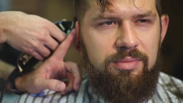 Creando un nuevo look de cabello. joven barbudo hombre consiguiendo corte de pelo por peluquería mientras sentado en silla en barbería — Vídeos de Stock