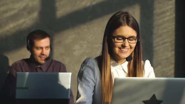 Smart giovane call center esecutivo femminile sorridente a voi in un ambiente luminoso ufficio con collega maschile sullo sfondo — Video Stock