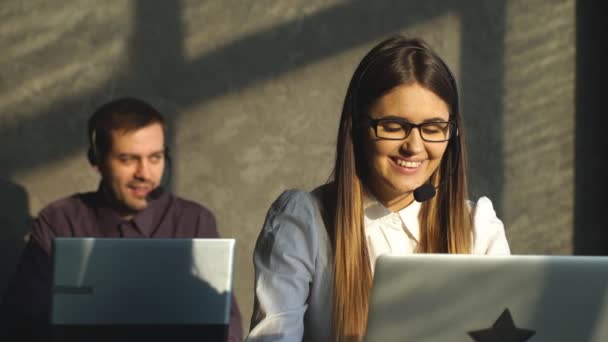 Inteligente joven centro de llamadas ejecutiva femenina sonriendo a usted en un ambiente de oficina brillante con colega masculino en el fondo — Vídeos de Stock