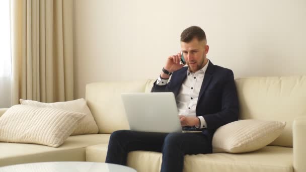 Tetap berhubungan dengan rekan-rekan. Pemuda tampan berbaju sedang mengerjakan laptop dan berbicara di telepon genggam — Stok Video
