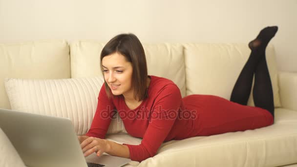 Atractiva mujer elegante acostada en el sofá de la habitación del hotel y trabajando en el ordenador portátil . — Vídeos de Stock