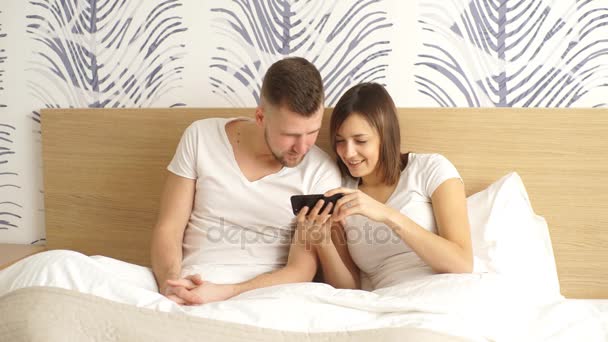 Ευτυχισμένο ζευγάρι αγκαλιά στο κρεβάτι με smartphone στο σπίτι στην κρεβατοκάμαρα — Αρχείο Βίντεο