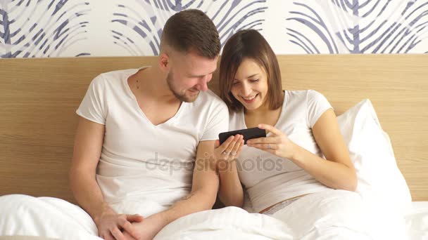 Bonito casal feliz jovem na cama com o telefone móvel sorrindo. Ambiente doméstico, estilo de vida foto . — Vídeo de Stock