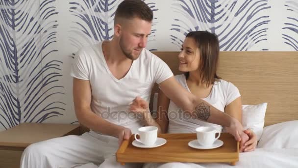 Młody człowiek przyniósł kawy w łóżku podczas jego dziewczyna siedzi na łóżku ubrana piżamy w sypialni — Wideo stockowe