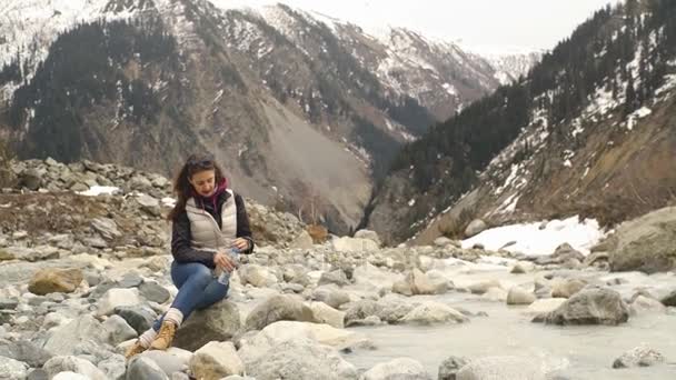 Hermosa mujer viajero bebe agua cerca de un río de montaña en un fondo de montañas — Vídeos de Stock