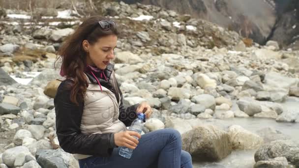 Beautiful woman traveler drinks water near a mountain river on a background of mountains — Stock Video