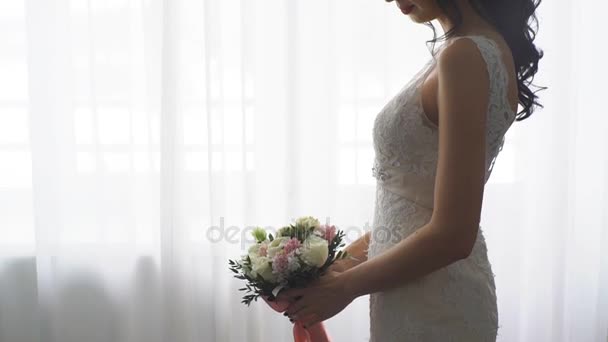 Брюнетка невеста стоит у окна и нюхает букет невесты — стоковое видео