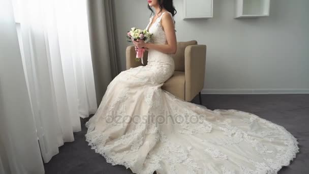 Encantador jovem noiva está sentado em poltrona em um vestido de noiva com um grande buquê de flores, sonhando acordado — Vídeo de Stock