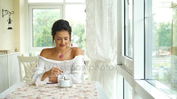 Hermosa morena en una blusa blanca, bebiendo café en la terraza luminosa — Vídeo de stock