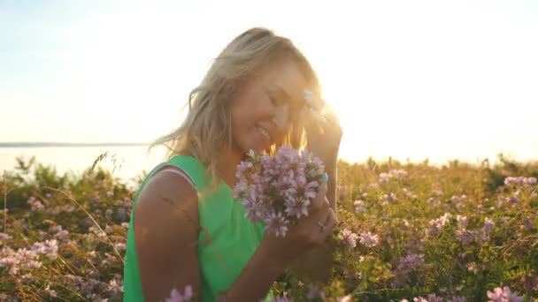 Schöne Blondine in einem hellgrünen Kleid sammelt wilde Blumen und posiert bei Sonnenuntergang für die Kamera — Stockvideo