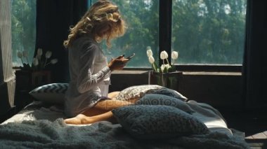 genç güzel sarışın pencereden yatakta oturuyor ve telefonda bir ileti yazdırır
