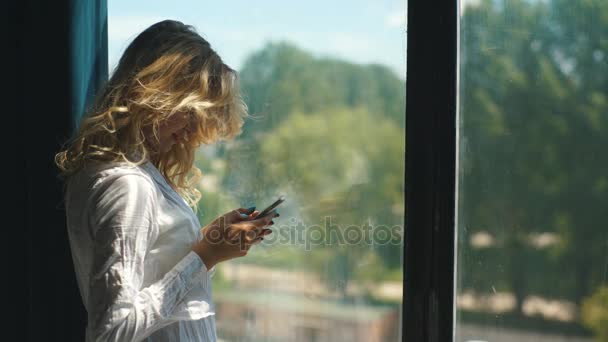 Jovem bela loira na janela e ler uma mensagem no telefone — Vídeo de Stock