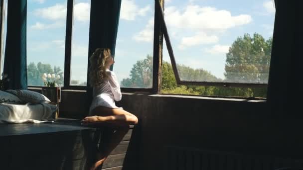 Mooie sexy blonde vrouw in wit shirt en ondergoed poseren voor de camera in de buurt van het venster — Stockvideo