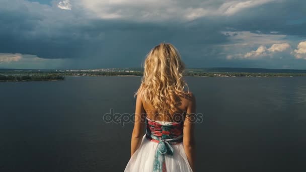 La blonde lève les bras au bord d'une falaise, une rivière en arrière-plan, vue arrière — Video