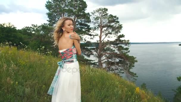 Rubia en un hermoso vestido de pie en el borde del acantilado y mirando al río — Vídeos de Stock