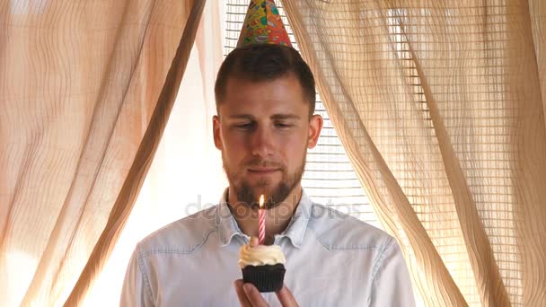 Een man in een feest hoed blaast de kaars op de cupcake — Stockvideo