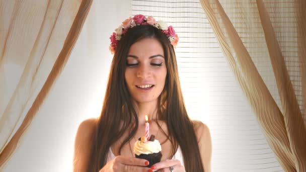 Een jonge brunette vrouw in een feest hoed blaast de kaars op de cupcake — Stockvideo