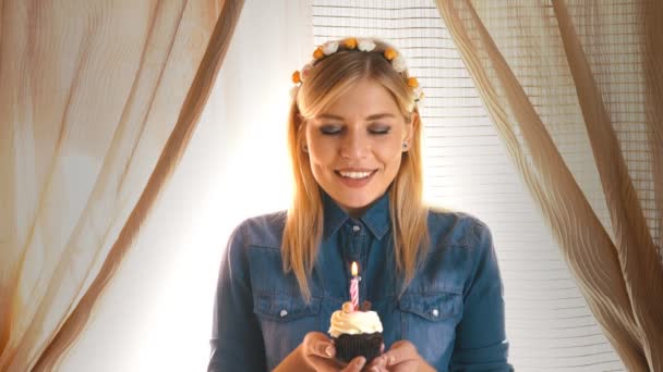 Μια νεαρή ξανθιά γυναίκα σε ένα καπέλο κόμμα φυσάει κερί σε cupcake — Αρχείο Βίντεο