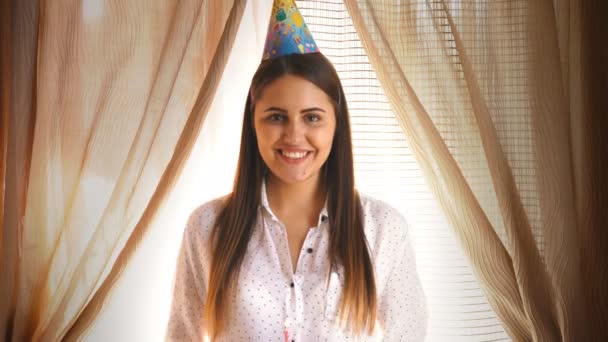 Una giovane donna bruna in un cappello da festa spegne la candela su un cupcake — Video Stock