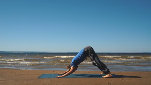 Jovem homem de pé na posição Downward Facing Dog na praia — Vídeo de Stock