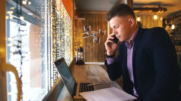 Молодой бизнесмен разговаривает по телефону и работает за ноутбуком в кафе — стоковое видео