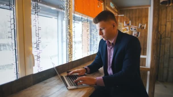 Lavora al bar. Giovane allegro in abbigliamento casual intelligente guardando il computer portatile e seduto vicino alla finestra in ufficio creativo o caffè — Video Stock