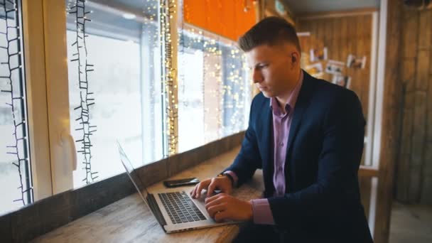 Zelfverzekerde jonge man in vrijetijdskleding, werk op laptop terwijl u zit in de buurt van venster in creatieve kantoor of café — Stockvideo