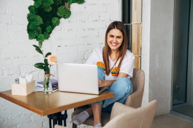 bir dizüstü bilgisayar ile kafede çalışan ve kameraya gülümseyen serbest meslek sahibi bir genç kızdır