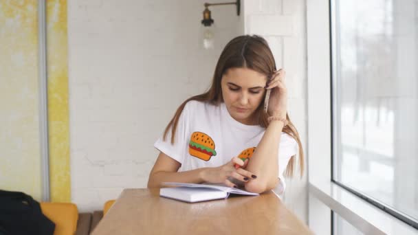 Joven mujer de negocios sentada cerca de la ventana, habla por teléfono y registra datos en un cuaderno — Vídeos de Stock