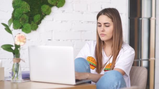 Una giovane ragazza è una freelance che lavora nel caffè con un computer portatile e sorride alla macchina fotografica — Video Stock