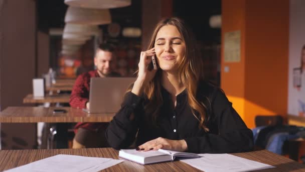 Vidám fiatal gyönyörű nő ül egy kávézóban, és beszél a telefonon — Stock videók
