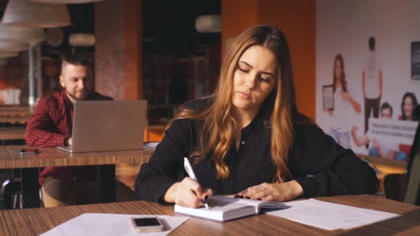 Chica joven de negocios escribiendo nota en el cuaderno en la cafetería, hombre con el ordenador portátil en el fondo — Vídeos de Stock