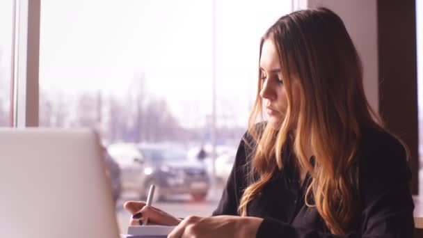 Mulher de negócios séria em uma camisa preta que trabalha em um laptop e toma notas em um caderno — Vídeo de Stock