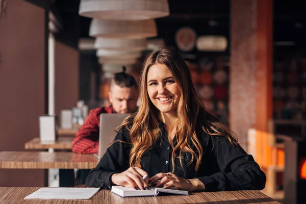 Mujer joven sonriendo a la cámara mientras trabajaba remotamente en la cafetería. Mujer positiva con bloc de notas —  Fotos de Stock