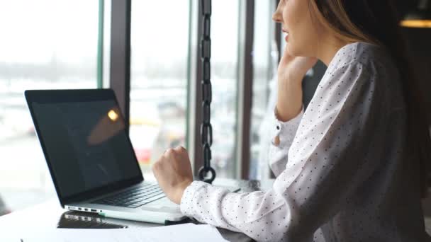 Ung affärskvinna klädd i vit skjorta sitter på café på träbord framför laptop och talar i mobiltelefon. — Stockvideo