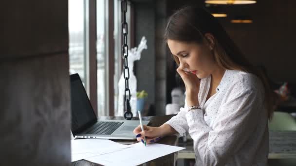 Молодая предпринимательница работает в кафе с ноутбуком, подписывает документы и разговаривает по телефону — стоковое видео