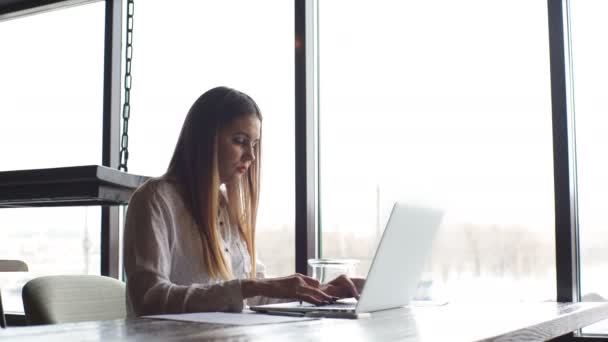 Бізнес-леді, що друкує на ноутбуці, сидить у кафе біля вікна — стокове відео