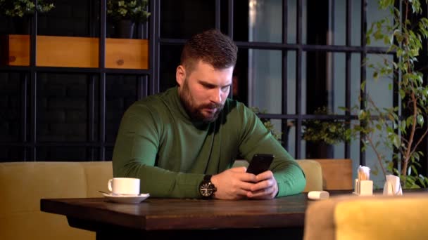 Joven barbudo hipster hombre se sienta a la mesa en la cafetería, utiliza un teléfono inteligente y beber café — Vídeos de Stock