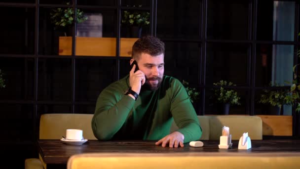 Joven barbudo hombre alegre sentado en la cafetería en la mesa, trabajando en la computadora, hablando por teléfono móvil — Vídeos de Stock