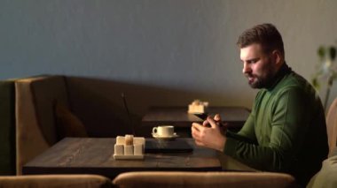 Sakallı hippi işadamı, kafeteryada oturuyor, laptopta çalışıyor, cep telefonuyla konuşuyor..