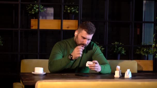 Um homem hipster senta-se em um café e ouve música em fones de ouvido Bluetooth — Vídeo de Stock