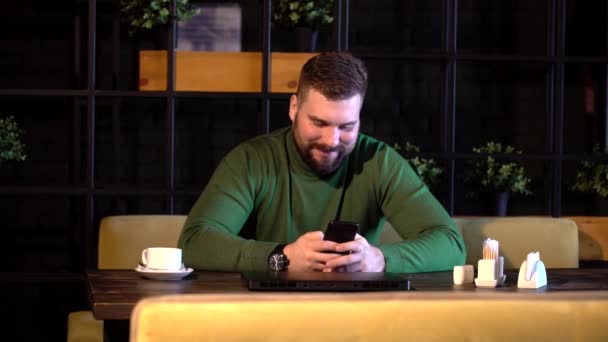 Hombre barbudo feliz sentado en un café y usando un teléfono inteligente — Vídeo de stock