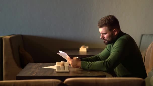 Joven atractivo barbudo hipster hombre se sienta en la cafetería leyendo documentos en papel — Vídeos de Stock