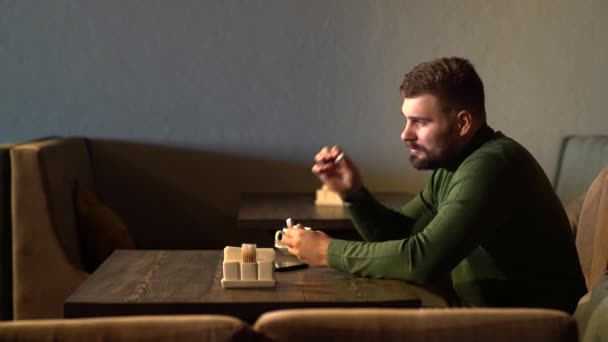 一个嬉皮士男人坐在咖啡店里，用蓝牙耳机听音乐 — 图库视频影像