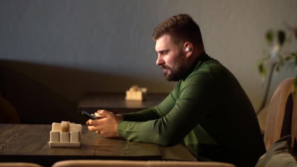 Joven hombre de negocios guapo está trabajando en un ordenador portátil en una cafetería y hablando en un auricular inalámbrico — Vídeo de stock